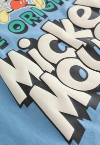 Gros plan sur un tissu bleu avec un grand texte blanc et noir en gras "Mickey Mouse" et un personnage de dessin animé partiel en rouge, jaune et noir.