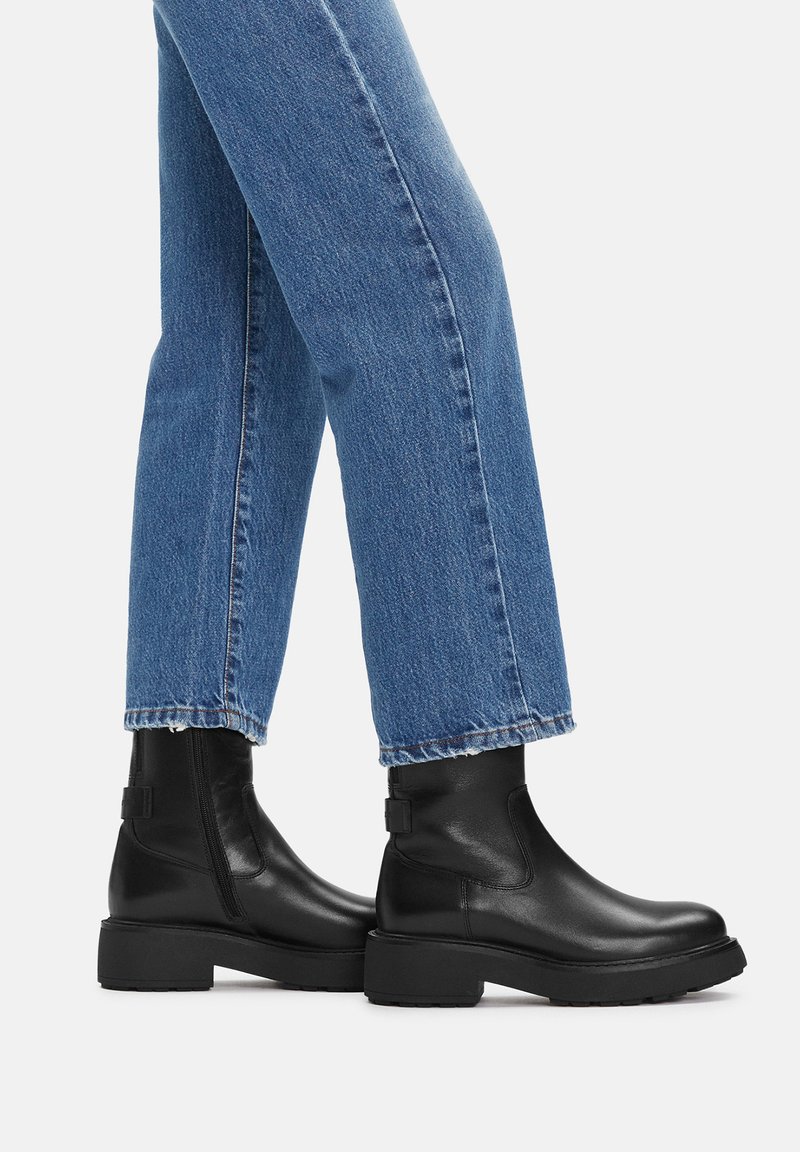 Czarne skórzane botki za kostkę o matowym wykończeniu, okrągłym nosku i masywnej podeszwie. W połączeniu z niebieskimi dżinsami prostymi.