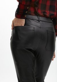 Pantaloni in ecopelle neri con una texture liscia, dotati di due tasche posteriori e una vestibilità slim, abbinati a una blusa a quadri.