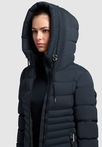 Mornarska puffer jakna s velikom kapuljačom, kvadriranim uzorkom, zatvaranjem na zatvarač i vezicama. Ima brendirani akcent na rukavu.