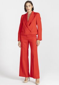Czerwony garnitur na miarę, składający się z krótkiego blezera na dwa guziki oraz spodni o szerokich nogawkach. Wykonany z gładkiej tkaniny o eleganckim wykończeniu.
