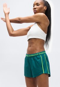 Γυναίκα με λευκό αθλητικό σουτιέν και πράσινα σορτς Under Armour τεντώνει το χέρι της μπροστά από το στήθος της, με φόντο μια μονόχρωμη επιφάνεια.