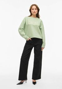 Felpa verde menta con la scritta "CHÉRIE" in testo crema, abbinata a pantaloni neri a gamba larga. Il modello indossa tacchi neri appuntiti. Texture liscia.