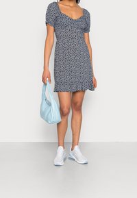 Mörkblå klänning med blommönster, puffärmar och volangkant. Matchad med en ljusblå handväska och vita sneakers, vilket framhäver en avslappnad look.