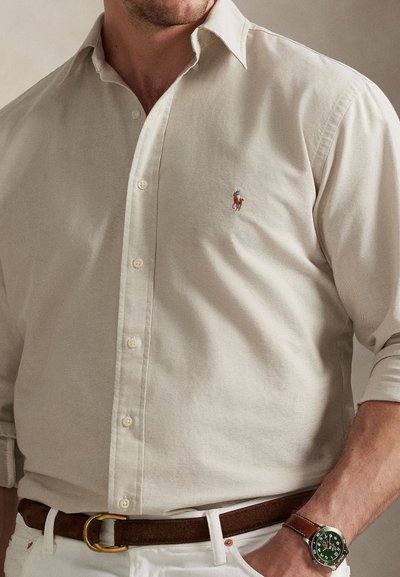 Beige skjorte med knapper og krage, med en liten brodert logo. Kombinert med et brunt belte og armbåndsur, med en glatt tekstur.