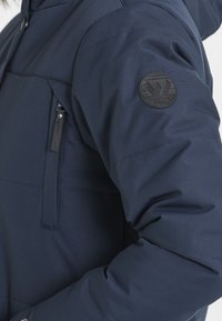 Marinblå isolerad jacka med en ficka på bröstet som stängs med dragkedja och en rund svart logga på vänster ärm, tillverkad av ett slätt, strukturerat material.