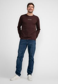 Czerwony t-shirt z długim rękawem z logo "PETROL", w połączeniu z ciemnoniebieskimi dżinsami i białymi trampkami, na jednolitym białym tle.