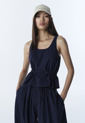 Navyblauw mouwloos topje met een geplooide taille en trekkoorden, gecombineerd met losse broeken met zijzakken. Gecombineerd met een lichtbeige pet.