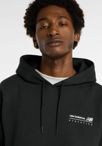 Haut à capuche noir en mélange de coton, avec une capuche à cordon et un logo blanc "New Balance Athletics" sur la poitrine.