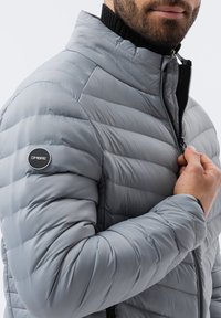 Jasnoszara pikowana kurtka z wype�łnieniem, z poziomym przeszyciem, wysokim kołnierzem i czarnym zamkiem błyskawicznym. Na lewym ramieniu znajduje się okrągłe logo.