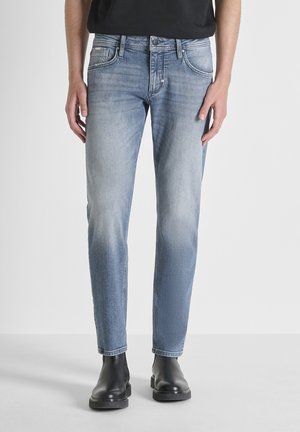 Uomo che indossa jeans skinny azzurri chiari e stivaletti in pelle nera, in piedi contro uno sfondo bianco uniforme.