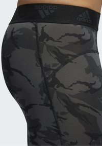 Zbliżenie na legginsy w czarno-szary wzór camo z widocznym szwem i logo Adidas na czarnej elastycznej talii.