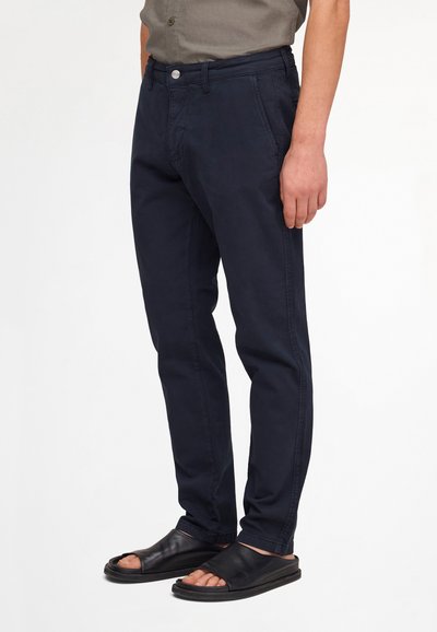 Homme portant un pantalon bleu marine foncé et des sandales en cuir noires à bout ouvert, debout devant un fond uni.