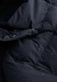 Tela de chaqueta impermeable en color azul marino con superficie texturada, gotas de agua sobre el material, con compartimentos cosidos y un broche negro.