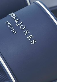 Κοντινό πλάνο σε σαγιονάρα Jack & Jones Studio σκούρου μπλε με λευκό ανάγλυφο όνομα της μάρκας στο λουράκι και λευκή διακοσμητική λεπτομέρεια.