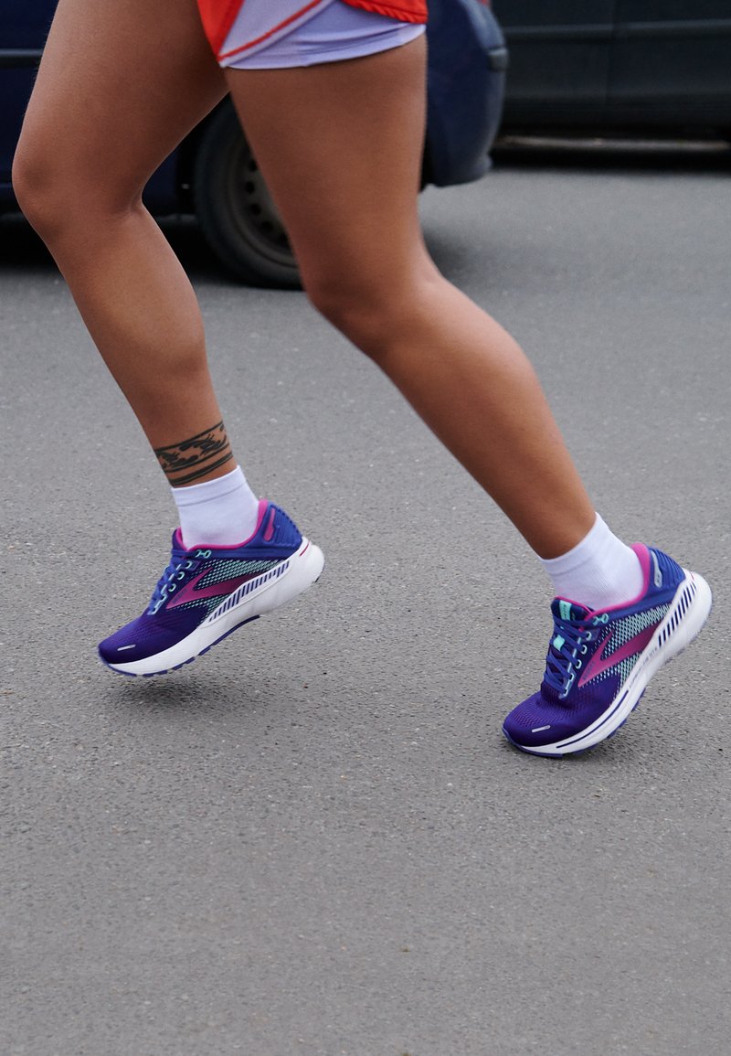 Zapatillas deportivas moradas con acentos azules, parte superior de malla transpirable, suela blanca y cordones decorativos, que se llevan con calcetines blancos mientras se camina.