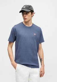 Νεαρός άνδρας που φοράει μπλε κοντομάνικο μπλουζάκι, λευκό παντελόνι και σκούρο γκρι καπέλο, με τατουάζ στο αριστερό του αντιβράχιο.