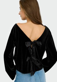 Top lungo a maniche, in velluto nero, con un profondo scollo a V sul retro, arricchito da un dettaglio a fiocco. Abbinato a jeans in denim azzurro chiaro.