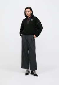 Sort fleecepullover med halv lynlås og "GAP"-logo, kombineret med højtaljede grå vidde-bukser og sorte spidse sko.