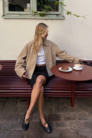 Beige zip-jakke med overdimensionerede ærmer, hvid top, sorte shorts og sorte flade sko. Bord med kaffe og kage, placeret mod en neutral væg.