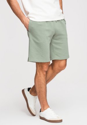 Homme portant un short décontracté vert clair, un T-shirt blanc et des baskets blanches avec des semelles marron, debout les mains dans les poches.