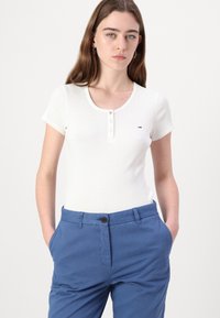 White ribbed t-shirt with a round neckline, short sleeves, and button detail. Paired with blue cotton pants featuring a button closure.