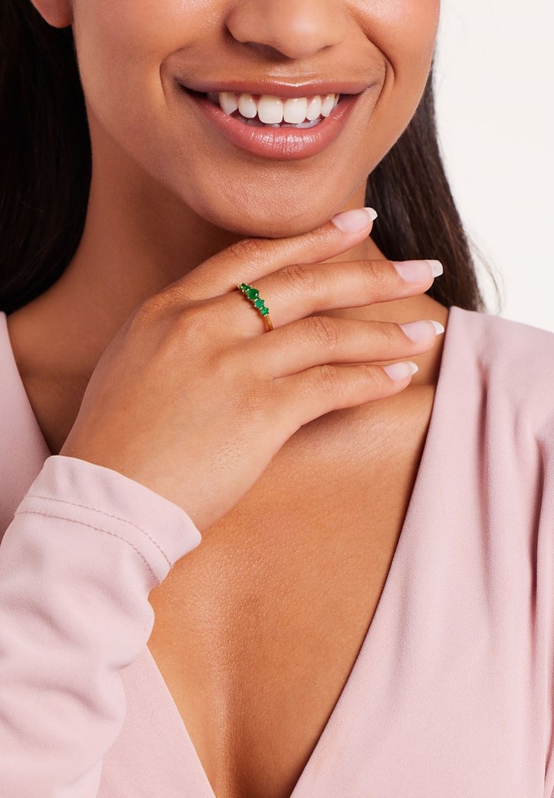 Gold ring featuring three green gemstones, worn on a hand with a natural nail finish, against a light pink shirt.