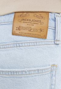 Lichtblauwe denim jeans met een tan leren label met "JACK & JONES" erop en stiksels langs de tailleband en zakken. Kenmerk van de Heritage-collectie zichtbaar.