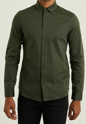 Hombre con camisa ajustada de manga larga color verde oscuro con botones y cuello, y pantalones negros, de pie con los brazos relajados.
