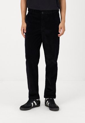 Pantalons en velours côtelé noir avec une coupe droite, dotés de côtes verticales, de poches avant et d'une fermeture à bouton. Associés à des baskets noires et blanches.