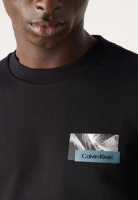 Zwarte katoenen T-shirt met een ronde hals, voorzien van een grafische patch met een zwart-wit ontwerp en "Calvin Klein" in blauwe tekst.