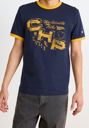 Homme portant un t-shirt bleu marine avec une bordure jaune et un grand texte jaune usé ainsi que des graphismes indiquant "CHP Worldwide Tribe" sur le devant.
