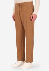 Pantaloni in pile marrone con vita regolabile, dettagli a pieghe e tasche laterali, abbinati a sneakers bianche su uno sfondo neutro.