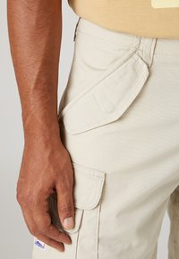 Pantaloni cargo beige con tasca laterale, realizzati in tessuto leggero. Presentano cuciture distintive e un piccolo logo del marchio in basso.