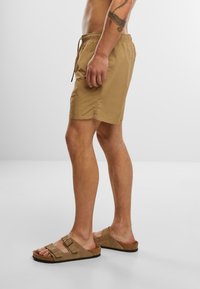 Pantaloni scurti de baie bej cu talie elastică și șnur, asortați cu sandale slip-on din suede maro, având două accente cu cataramă.