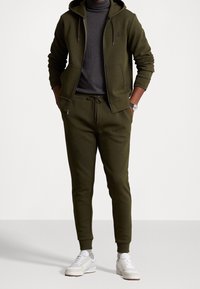 Sudadera verde oliva con cremallera frontal, cordón y un logo; combinada con joggers a juego y zapatillas blancas. Material de mezcla de algodón.