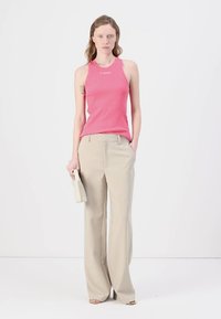 Mønstret rosa ribbet tanktop med logo, kombinert med lysebeige vide bukser. Modellen holder en liten hvit håndveske. Enkel bakgrunn.