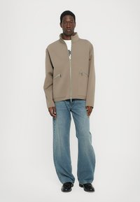 Chaqueta de cremallera color marrón claro con cuello alto, dos bolsillos frontales con cremallera, combinada con jeans azules holgados y zapatos negros. Textura suave.