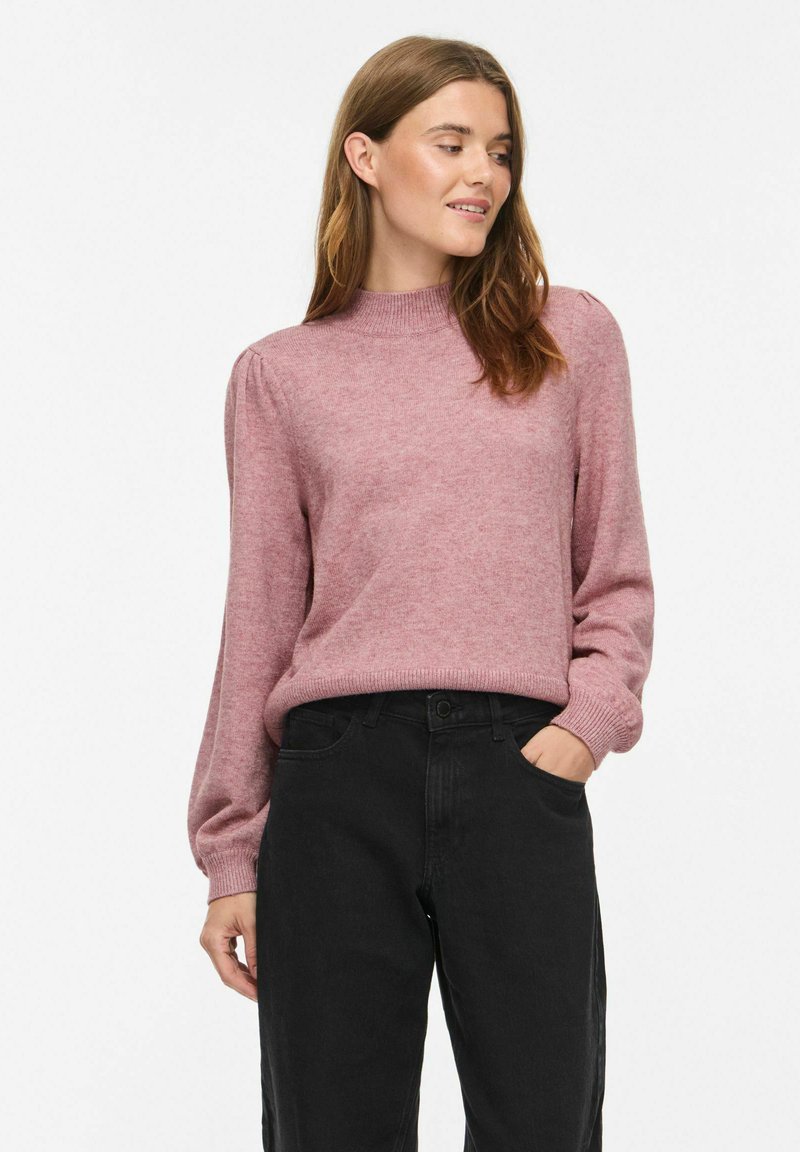 Femme portant un pull rose à manches longues et un pantalon noir, debout avec une main dans la poche, regardant sur le côté sur fond blanc.