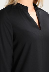 Schwarze Bluse mit einem Rüschenkragen und einem V-Ausschnitt. Hergestellt aus glattem Stoff mit langen Ärmeln. Minimalistische und elegante Gestaltung.