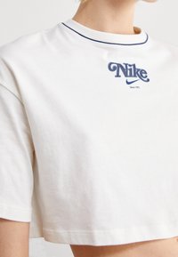 Vit bomullstopp med kort ärm som har en marinblå broderad "Nike"-logotyp med rundad text och en kontrasterande söm vid halsringningen.