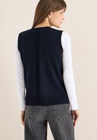 Granatowa dzianinowa kamizelka z prążkowaną fakturą na plecach. Noszona na białej koszuli z długim rękawem, zestawiona z szarymi jeansami.