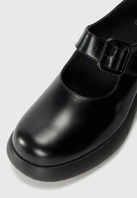 Black leather Mary Jane shoe with a rounded toe, a buckle strap, and a textured black rubber sole. Smooth finish with an enclosed design.