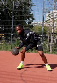 Svart atletisk jakke med hvite striper, svarte shorts og lyse gule joggesko. Holder en basketball på en rød utendørs basketballbane med et gjerde.