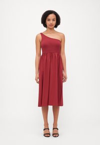 Vestido burdeos de un solo hombro hecho de una tela suave; el cuerpo ajustado fluye hacia una falda fruncida. Llevado con sandalias negras de tiras.