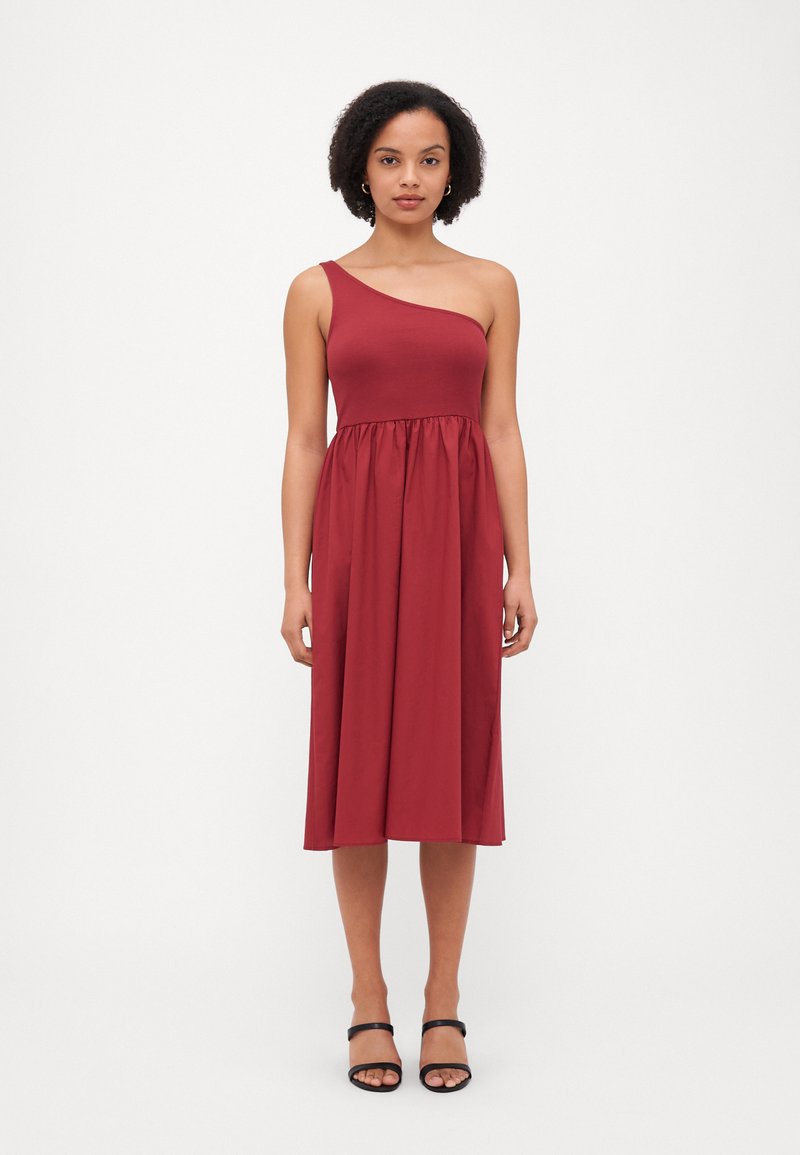 Vestido burdeos de un solo hombro hecho de una tela suave; el cuerpo ajustado fluye hacia una falda fruncida. Llevado con sandalias negras de tiras.