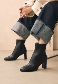 Schwarze Leder-Stiefeletten mit spitzer Kappe und klobigem Absatz. Das Design verfügt über einen seitlichen Reißverschluss und eine glatte Oberfläche. Jeanshosen mit umgeschlagenem Saum.