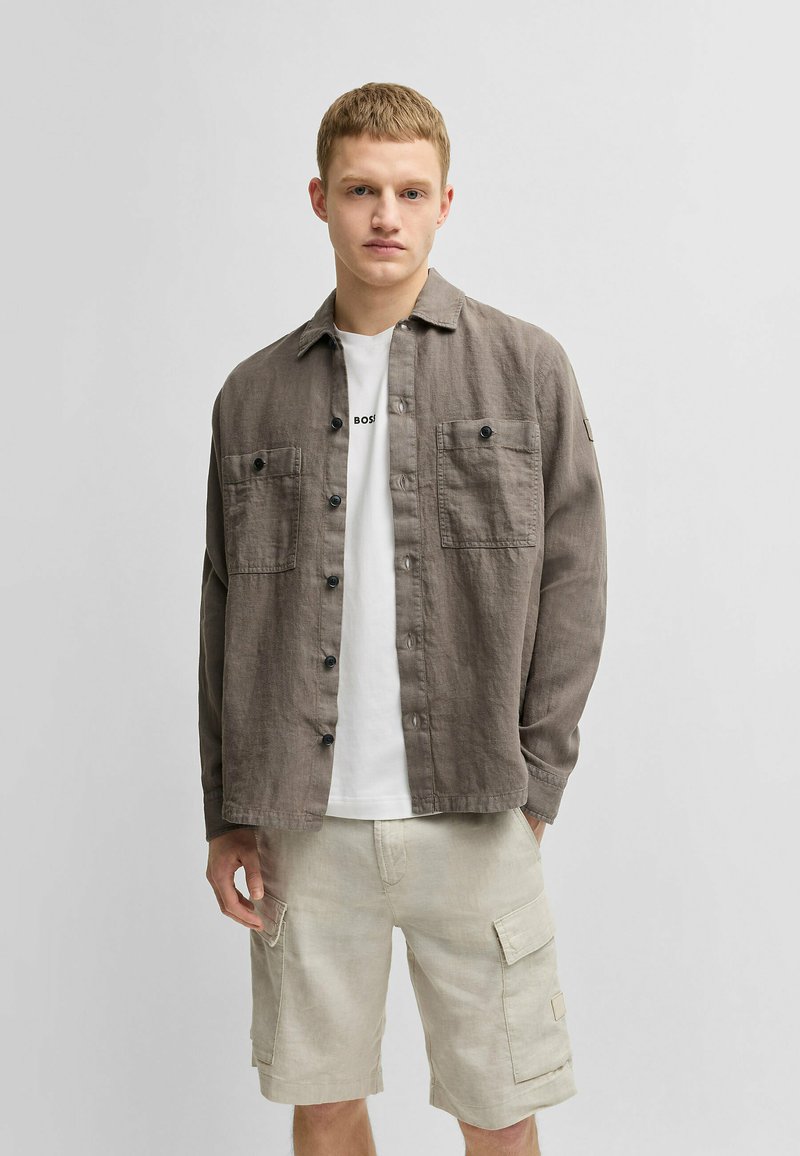 Lichtbruine linnen shirt met twee borstzakken en zwarte knopen, gedragen over een wit T-shirt en gecombineerd met lichtbeige cargoshorts.