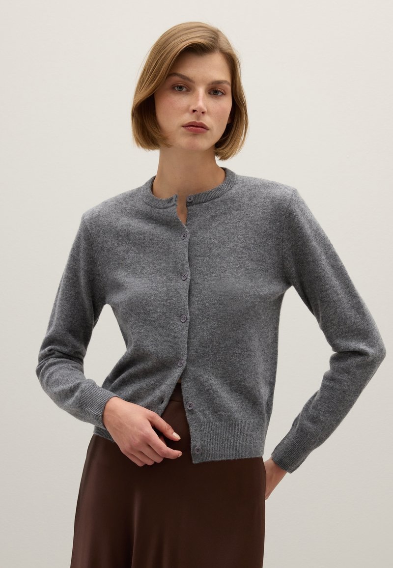 Cardigan grigio a maglia con collo rotondo, bottoni frontali, vestibilità slim e polsini a coste. Indossato sopra un vestito marrone, con una texture di tessuto liscia.
