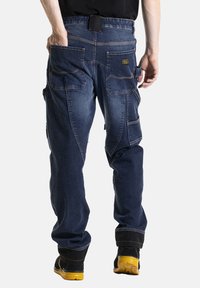 Pantalons cargo en denim bleu avec une coupe décontractée, dotés de plusieurs poches, de coutures contrastées et de zones de genoux renforcées. Chaussures noires visibles.